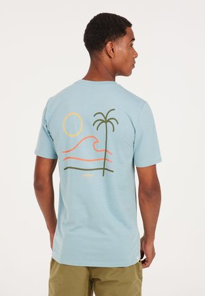 Camiseta de algodón azul claro con mangas cortas. En la parte posterior presenta un gráfico de olas, un sol y una palmera en naranja y verde. "PROTESTA" impreso debajo.