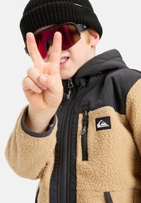 Chaqueta de forro polar beige con detalles negros, bolsillos con cremallera y un parche de logo. Gorro de lana negro y gafas de sol grandes con tintado rosa. Mano haciendo un signo de paz.