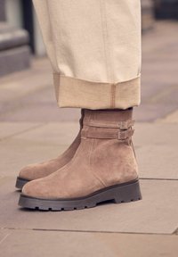 Beige mokasíny do členkov s okrúhlou špičkou a robustnou podrážkou s dezénom. Obsahujú dva pracky na boku pre bezpečné upevnenie.