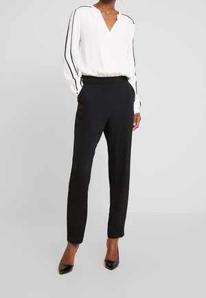 Personne portant un chemisier blanc à rayures noires sur les manches, un pantalon noir taille haute et des escarpins noirs pointus, debout devant un fond uni.