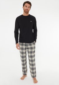 Chemise noire à manches longues associée à un pantalon de pyjama à carreaux beige et noir. La chemise présente un petit logo sur la poitrine ; texture de tissu douce.