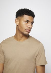 T-shirt de algodão bege com gola redonda, mangas curtas e uma textura suave. Ajusta-se de forma justa ao corpo, design minimalista sem padrões.