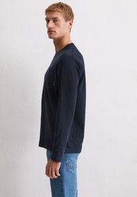 Granatowa koszula z długim rękawem, okrągłym dekoltem i kieszenią. Miękka w dotyku, o luźnym kroju. W zestawieniu z jasnoniebieskimi dżinsami.