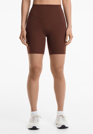 Bruine hoge taille fietsbroeken gemaakt van rekbaar, glad materiaal, die net boven de knie eindigen. Gecombineerd met witte sportschoenen.