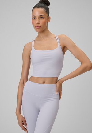 Kobieta o ciemnych włosach w kok, ubrana w jasnofioletowy krótki top i legginsy, pozująca z jedną ręką na biodrze na szarym tle.