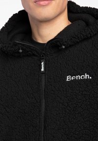 Nahaufnahme einer Person, die einen schwarzen, strukturierten Fleece-Kapuzenpullover mit dem auf der Brust aufgestickten Bench-Logo und Reißverschlussanhänger trägt.
