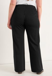 Vrouw in zwarte gestreepte wijde broek, gezien van achteren, staand tegen een eenvoudige lichte achtergrond.