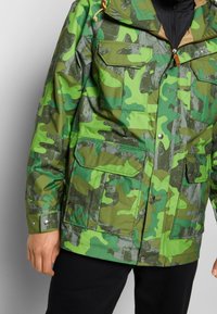 Camouflage jacket in various shades of green. Features a zip front, two chest pockets, and adjustable drawstring hood. Smooth, waterproof material.