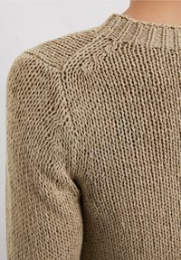 Gros plan sur un pull en tricot beige montrant des motifs détaillés de côtes et de points torsadés sur l'épaule et le haut du bras.