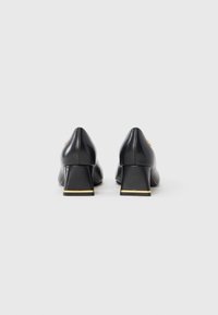 Black leather heels featuring a pointed toe and a chunky heel with a gold accent at the base. Smooth texture with minimalist design.