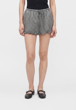 Kvinde iført sort/hvide ternede shorts med snøre, sort top og sorte flade Mary Jane sko, stående mod en ensfarvet baggrund.