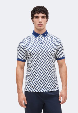 Mand iført en kortærmet, blå og hvid mønstret polo-shirt med en mørkeblå krave og mørkeblå bukser, stående mod en ensfarvet lys baggrund.