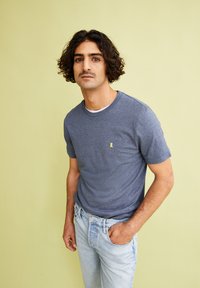 T-shirt a maniche corte blu navy con colletto rotondo e logo in nastro giallo. Indossato con jeans in denim azzurro. Tessuto morbido, vestibilità casual.