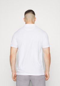 Mies, jolla on lyhyet hiukset, pukeutuneena valkoiseen lyhythihaiseen poolopaitaan ja vaaleanharmaisiin housuihin, nähtynä selkäpuolelta yksinkertaista valkoista taustaa vasten.