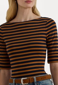 Ženska oblečena v kratkoslečeno majico s horizontalnimi rjavimi in črnimi črtami, vpeti v modre kavbojke z rjavim nubuk pasom.