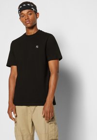 Black cotton t-shirt with short sleeves, featuring a small white logo. Model wears beige cargo shorts with side pockets.