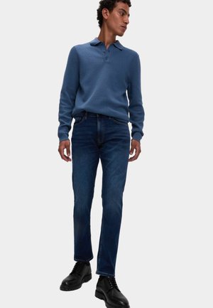 Niebieski, teksturowany sweter z kołnierzem polo, zestawiony z ciemnoniebieskimi jeansami. Noszony z czarnymi, sznurowanymi butami. Model stoi w profilu.
