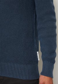 Marineblauer Strickpullover aus strukturiertem Material, mit gerippten Bündchen und Saum; hat ein kleines Etikett auf der linken Seite, nahe dem unteren Ende.