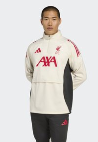 Ung mand iført en cremet og sort Liverpool FC-jakke med røde AXA- og Adidas-logoer, som står foran en ensfarvet lys baggrund.