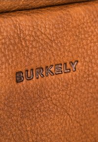 Cuero marrón texturizado con grano visible; logo "BURKELY" en relieve en marrón oscuro. El primer plano resalta los patrones naturales del material.