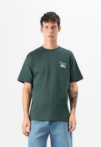 Young man wearing a dark green "Diavola" t-shirt with a graphic and light blue jeans, standing against a white background.