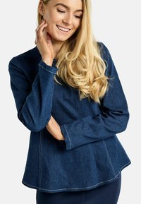 Temno modra denim bluza z dolgimi rokavi, zaobljenim robom in kontrastnimi belimi šivi. Mehka tekstura in priložnostna zasnova.