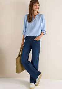 Lyseblå, langærmet bluse med V-hals; mørkeblå vide jeans; cremefarvede sneakers med blå detaljer; olivengrøn tote-bag.