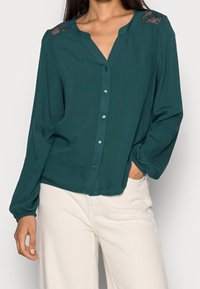 Blouse boutonnée turquoise à manches longues, avec accents en dentelle sur les épaules, encolure ronde et coupe décontractée, associée à un pantalon en velours côtelé crème clair.