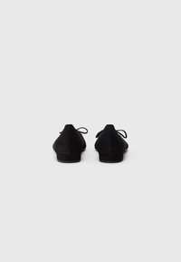 Svarte mokkasiner i semsket skinn med lav hel. Har en glatt overflate og båndsnøring bak. Minimalistisk design med spiss tåform.