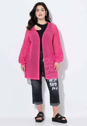 Cardigan a maglia aperta e maniche lunghe di un brillante colore rosa, indossato sopra un top nero con scollatura, abbinato a jeans neri strappati e sliders neri.