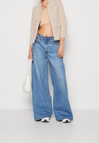 Chaqueta corta beige claro, top con borde en forma de ondas color canela y vaqueros anchos de mezclilla azul. Bolso blanco y zapatillas negras completan el conjunto.