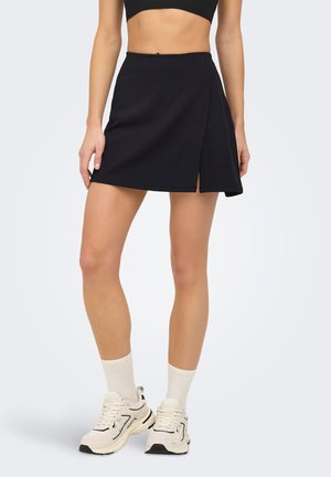 Kvinne iført svart omslagsstil mini-skjørt med liten splitt, hvite sportsokker og beige sneakers, stående mot en ensfarget bakgrunn.