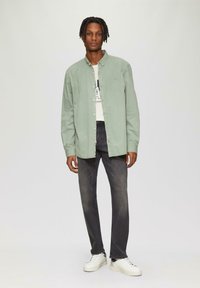 Jasnozielona koszula zapinana na guziki noszona na białej koszulce z grafiką, w połączeniu z ciemnymi dżinsami i białymi butami sportowymi. Model stoi na neutralnym tle.