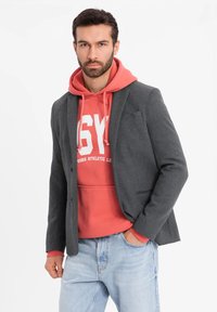 Grauer Blazer über einem korallenfarbenen Hoodie mit weißer Schrift, kombiniert mit hellblauen Jeans. Der Blazer hat eine taillierte Form und eine glatte Textur.