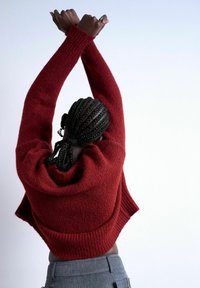 Personne avec des cheveux tressés, portant un pull rouge et un pantalon gris, étire les bras croisés au-dessus de la tête sur un fond uni clair.