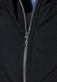 Chaqueta acolchada negra con cierre de cremallera que presenta una cremallera metálica. La tela muestra un patrón texturizado y acolchado, además de un diseño con capucha.