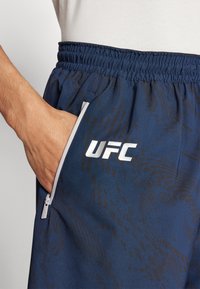 Mornarske kratke hlače z vrvico v pasu, stranskimi zadrgo žepi in subtilnim vzorcem. Na sprednji strani je natisnjena UFC logotip v beli barvi.