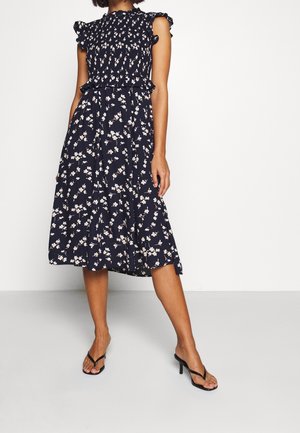 Femme portant une robe midi sans manches bleu marine à imprimé floral blanc avec des détails à volants, associée à des sandales à talons noires.
