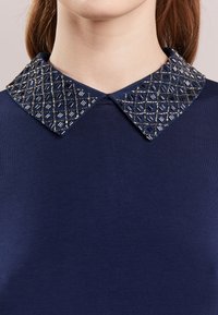 Top azul marino con un cuello texturizado y adornado, que presenta un patrón geométrico de hilos plateados y negros, enfatizando el contraste y el detalle.