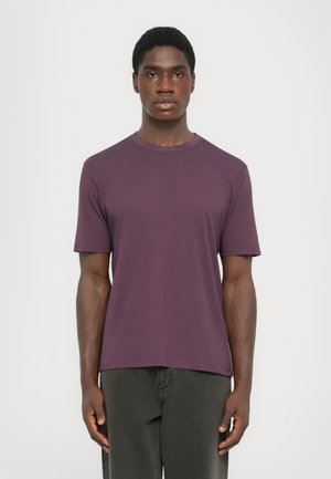 Young man with short hair stands facing forward wearing a plain purple short-sleeve t-shirt and dark grey pants against white background.
