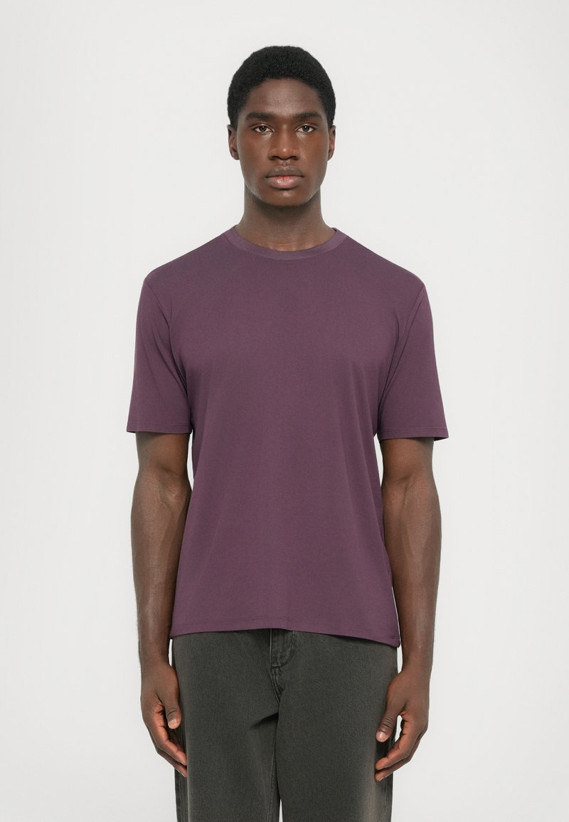 Young man with short hair stands facing forward wearing a plain purple short-sleeve t-shirt and dark grey pants against white background.