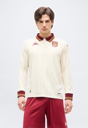 Joven con polo de manga larga color crema, con cuello y escudo en color granate y dorado, combinado con pantalones deportivos granates.