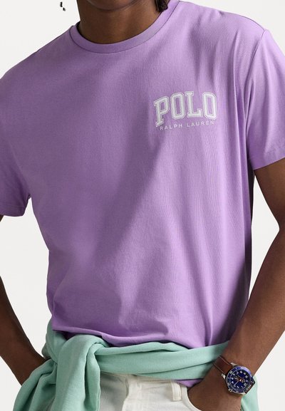 Lila pamut póló fehér "POLO RALPH LAUREN" logóval. mentazöld anyaggal a deréknál és kék órával a csuklón.