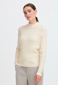 Maglione a collo alto lavorato a maglia in crema con un motivo testurizzato di cavi in rilievo. Silhouette aderente, maniche lunghe e polsini e orlo a coste.