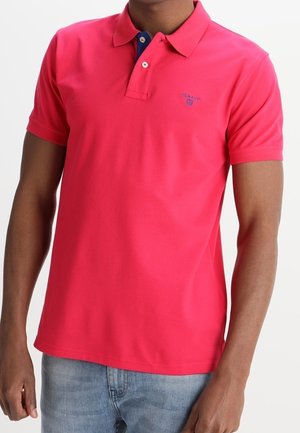 Man wearing bright red short-sleeve polo shirt with blue inner collar detail and light blue jeans, standing against white background.