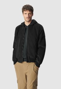 Czarna kurtka z kapturem, wykonana z miękkiego materiału, z zamkiem z przodu, prążkowanymi mankietami i luźnym krojem. Nosenia na ciemnej koszulce z dekoltem w serek i beżowych spodniach.