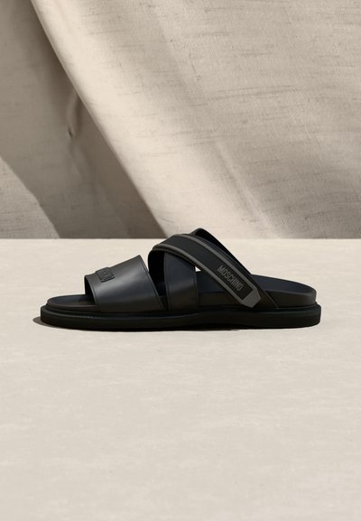 Sandale à glissière noire avec deux larges brides et une bride arrière ajustable ornée du logo "Moschino", posée sur une surface et un fond neutres.