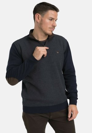 Homme portant un pull texturé bleu marine avec fermeture éclair et coudières, regardant sur le côté, une main près du col.