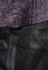 Fleece látka v tmavě fialové barvě nahoře, přecházející do černé síťoviny dole, s texturovaným povrchem a viditelným švem.