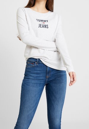 Woman wearing white long-sleeve Tommy Jeans shirt and blue denim jeans, standing with arms crossed against a plain light background.